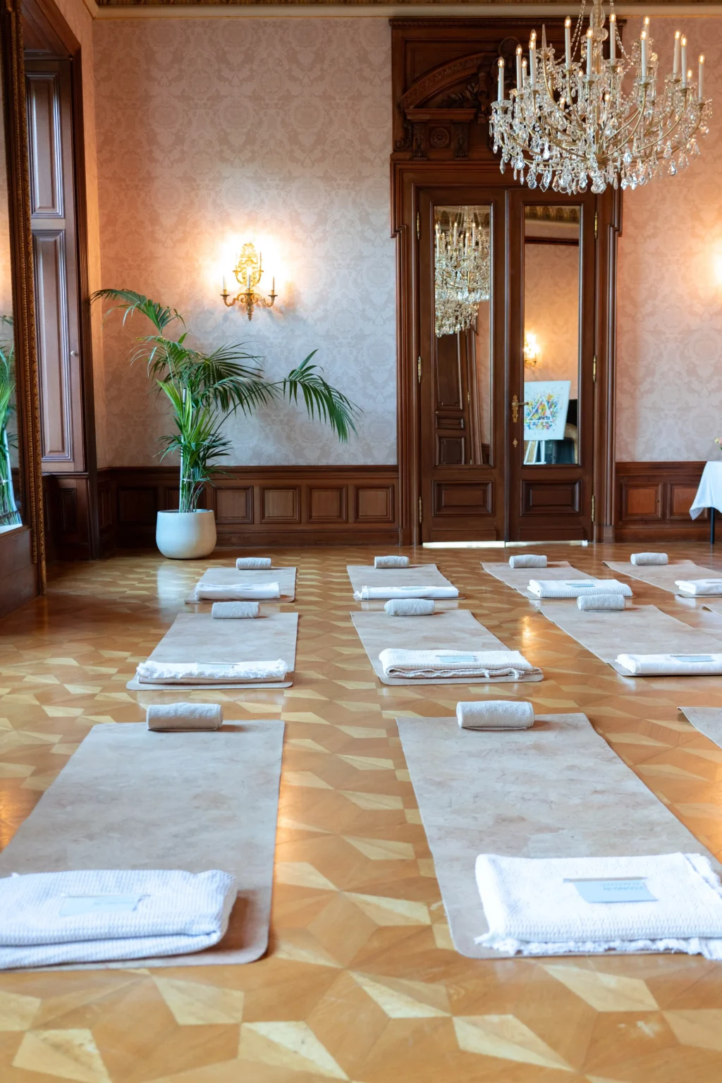 Ein Raum im Almanac Palais Vienna mit Kronleuchtern und großen Spiegeln, der für Yoga eingerichtet ist, mit Reihen von Matten und ordentlich aufgerollten Handtüchern auf einem gemusterten Holzboden.